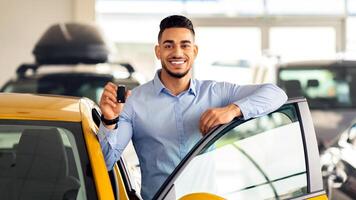 Handsome arab man customer holding key from auto and smiling, buying brand new car in showroom. Happy middle-eastern guy standing by nice yellow car and showing automatic key at camera photo