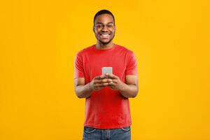 Happy African American Guy Using Phone With New Mobile Application Smiling To Camera Standing Over Yellow Background In Studio. People And Gadgets, Great App For Smartphone Concept photo