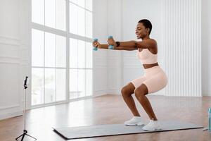 Positive sporty lady exercising and squatting with two dumbbells in front of cellphone on tripod at home, smiling fitness blogger recording for her followers having personal virtual training photo