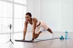 Online Training Concept. Sporty African American lady doing mountain climbers exercise, watching tutorial or having online class using cellphone on tripod, looking at screen, exercising at home photo