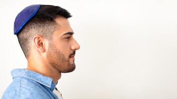 Male Portrait. Profile side view of serious jewish man standing and posing at studio, looking aside at free copy space, wearing blue kippa, posing isolated over white gray studio background, banner photo