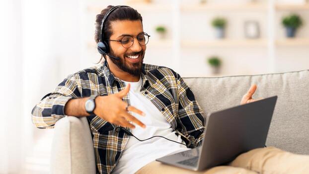 alegre indio chico reclinable en sofá en vivo habitación, utilizando ordenador portátil y inalámbrico auriculares, teniendo conferencia con cliente o Novia desde hogar, sonriente a cuaderno pantalla, gesticulando, Copiar espacio foto