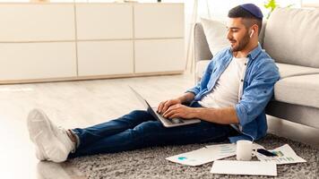 Workplace. Young Jewish Employee Working On Laptop Sitting On Floor Carpet, Busy Male Entrepreneur Typing On Keyboard And Looking At Computer Screen, Checking Email Doing Online Project, Free Space photo