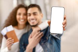 Happy Arab Couple Showing Smartphone With Blank Black Screen While Standing In Airport Terminal Together, Cheerful Middle Eastern Spouses Recommending New Avia Tickets Sales App, Mockup Image photo