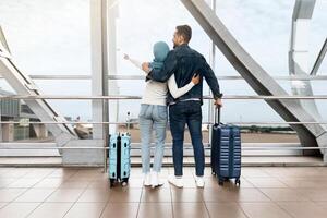 Weekend Travel. Loving Islamic Couple Standing Near Window In Airport And Pointing Away, Rear View Of Muslim Spouses With Suitcases Waiting For Flight At Terminal, Enjoying Upcoming Trip, Copy Space photo