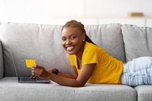 Online shopping, seasonal sale, black Friday concept. Smiling young black lady making payment, ordering goods on web, using internet bank services, lying on couch with laptop computer and credit card photo