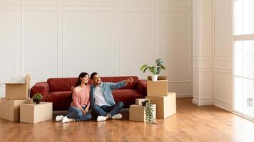 Portrait of smiling family sitting on the floor leaning on couch in new empty apartment. Guy pointing finger aside at free space embracing wife, choosing wall color and interior design repair, banner photo