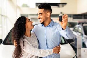 Loving arabic couple standing next to luxury white car and hugging at dealership automobile salon, showing key from their new auto, cheerfully smiling at each other and hugging, closeup photo