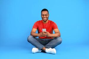 Happy Black Man Using Mobile Phone Browsing Internet Sitting Over Blue Background. Studio Shot Of Millennial Guy Using Social Media Application On Smartphone. New App For Your Phone Concept photo