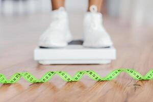 Weight Control Concept. Selective focus on yellow measuring tape on the floor and blurred view of african woman standing on scales, checking result of her slimming diet. Dieting, healthy living photo