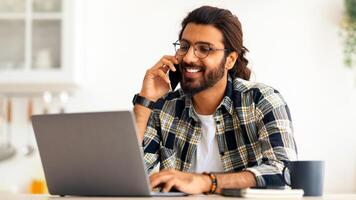 Smiling young arab man freelancer talking on mobile phone and taking notes, working from home during isolation, copy space. Happy guy having conversation on phone, using laptop at kitchen photo