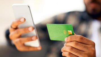 Green credit card and modern mobile phone in male hands, closeup photo. Unrecognizable middle-eastern man using cellphone, shopping online or banking while staying home, making order on Internet photo