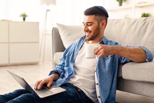 personas y tecnología concepto. sonriente joven judío hombre en kippa utilizando ordenador portátil participación taza y Bebiendo caliente café. masculino sentado en piso propensión en sofá acecho , película o seminario web, tomando descanso foto