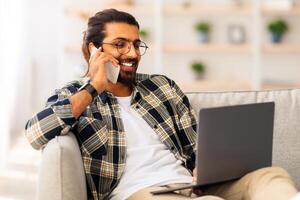 Positive long-haired, bearded arab guy freelancer sitting on sofa with lapto, having phone coversation, typing on computer keyboard, working from home, looking at screen and smiling, copy space photo