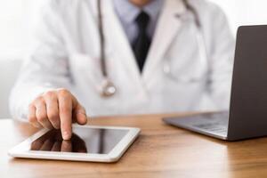 Close up of unrecognizable MD using tablet and computer, touching screen, sitting at wooden desk photo