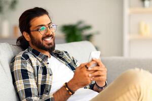 Handsome indian young man in casual relaxing on couch at home, playing games on smartphone, copy space. Cheerful arab guy using dating mobile application on celphone, chatting with ladies photo