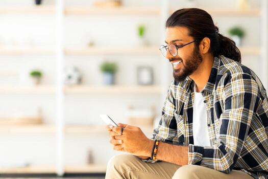 positivo de pelo largo Medio este chico con lentes sentado en sofá, utilizando Teléfono móvil, chateando con su Novia y sonriente, contento milenario árabe hombre Relajado a hogar, lado vista, Copiar espacio foto