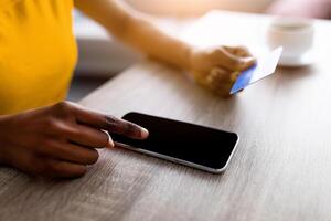 Unrecognizable black woman using smartphone and credit card for online shopping on table, closeup. Millennial lady ordering goods on mobile device, mockup for app or website design photo