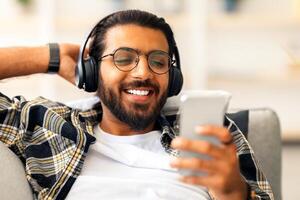 Closeup of cheerful arab guy watching movie while resting on couch at home, using wireless headset and mobile phone. Joyful middle-eastern young man freelancer relaxing on weekend or having break photo