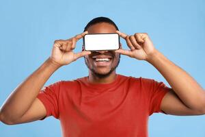 African American Guy Holding Smartphone With Empty Screen In Front Of Eyes Advertising Great Mobile Application On Blue Background In Studio. New App For Your Phone. Mockup photo