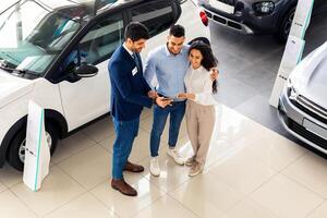 Top view of cheerful middle-eastern young couple having conversation with good-looking sales assistant at auto showroom, checking papers or car specification, full length photo