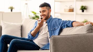 Phone Call, Mobile Communication And Voice Assistant Concept. Smiling Bearded Man In Kipa Talking On Smartphone Using Loudspeaker Mode, Guy Sitting On Comgortable Couch, Selective Focus photo