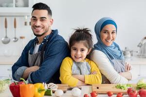 Portrait Of Happy Middle Eastern Mother, Father And Little Daughter Posing In Kitchen While Cooking, Confident Islamic Family Of Three Wearing Aprons Standing With Folded Arms And Smiling At Camera photo