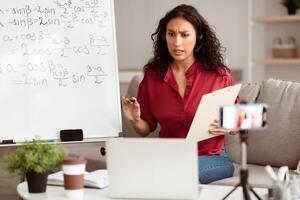 Online Studying, Distance Learning, Virtual Class. Serious focused lady teacher in wireless headset recording lesson on phone camera on tripod, sitting at desk near flipchart, talking to pc screen photo