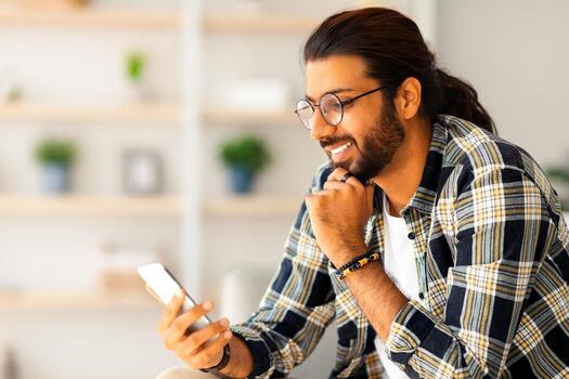 alegre de pelo largo Medio este chico con lentes sentado en sofá, utilizando teléfono inteligente, chateando con su Novia y sonriente, contento milenario árabe hombre Relajado a hogar, lado vista, Copiar espacio foto