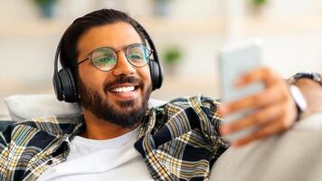 Handsome arab guy watching movie while resting on couch at home, using wireless headset and mobile phone, closeup. Joyful middle-eastern young man freelancer relaxing on weekend or having break photo