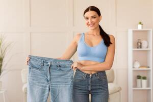 Slimming. Woman Comparing Jeans After Weight Loss Standing At Home, Smiling To Camera. photo
