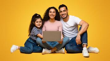 Portrait Of Happy Beautiful Arab Family Of Three With Laptop Computer Posing Together Over Yellow Studio Background, Cheerful Middle Eastern Parents And Their Little Daughter Smiling At Camera photo