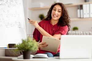 Young professional tutor recording tutorial for her blog using and looking at laptop screen, pointing pen at whiteboard with formulas. Online school, knowledge. Focus on teacher. E-learning photo