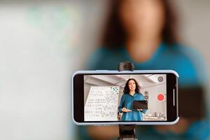 Modern Remote Education. Lady teacher shooting lecture on smartphone using tripod. Woman having conference with students, explaining material, live streaming. Selective focus on gadget screen photo