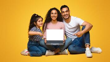 Cheerful arab family with little daughter showing laptop with blank white empty screen and pointing on it, happy middle eastern parents and female child recommending new website, mockup image photo