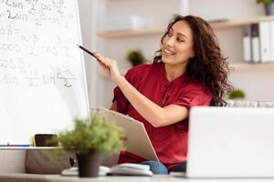 Portrait of smiling lady teacher chatting online, using laptop, tutor leading lesson, recording webinar, explaining, teaching students. Distance education, consulting from home, pointing on board photo