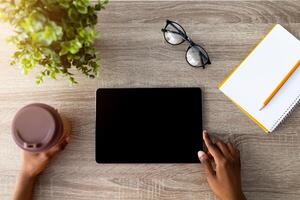 Top view of black woman with coffee using tablet computer for online shopping on wooden table, closeup of hands. Millennial lady ordering goods on touch pad with mockup for app or website design photo