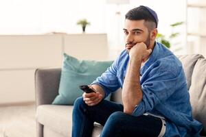 Boring Television Program. Bored Jewish Man Watching TV Switching Channels With Remote Control Sitting On Sofa At Home. Discontented Viewer Watching Uninteresting Movie, Feeling Lonely photo