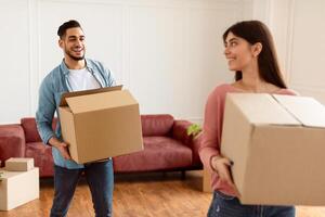 Loving couple carrying cardboard boxes together, helping each other in their new home on moving day. Young tenants relocating to new apartment with belongings, unpacking things in living room photo