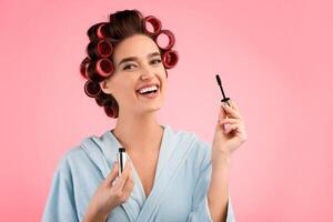 Eye Makeup. Happy Woman With Curlers Holding Mascara Applying It On Eyelashes Smiling To Camera Posing Wearing Dressing Gown Over Yellow Studio Background. Female Decorative Cosmetics photo