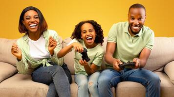 Domestic entertainment. Emotional black dad and daughter holding joysticks, competing with each other, parents playing games with child while sitting on sofa over yellow background photo