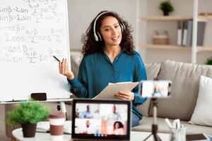 Online Training. Young lady tutor in headset teaching online by conference using laptop and smartphone. Teacher explaining information to diverse participants in the virtual conference photo