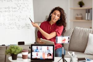 Online Lesson Concept. Portrait of young tutor pointing at board with trigonometric functions, explaining math to students making videochat. Woman in wireless headphones filming class on smartphone photo