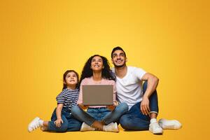 Portrait Of Smiling Young Arab Family Of Three With Laptop Looking Up At Copy Space Above Their Heads, Happy Middle Eastern Parents And Little Daughter Sitting On Floor Over Yellow Studio Background photo