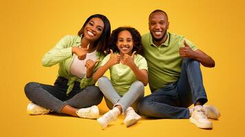 Positive african american family of three showing thumbs up gesture, approving or recommending something, sitting on floor at yellow studio background photo