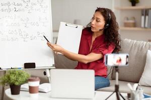 E-Learning. Confident woman having conference call using laptop. Lady teacher in headset showing clipboard, recording tutorial with mobile phone on tripod, streaming live pointing at board photo