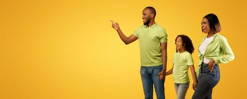 Positive african american family of three looking aside, young man pointing finger at copy space, standing on yellow background, demonstrating empty place for your advertisement, panorama photo