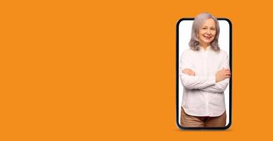 A senior woman with gray hair appears on a large smartphone screen, smiling while engaging in communication using a social media application. The background is bright and blank. photo