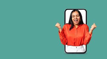 A young woman joyfully engages with a smartphone app showcasing her excitement. The background is plain, emphasizing her animated expression and the large screen of the phone. photo
