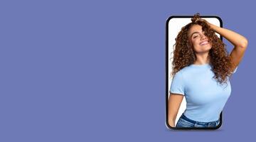 A cheerful young woman poses playfully with her smartphone, showcasing her curly hair and bright smile. She is engaged in social media communication while surrounded by a blank space. photo